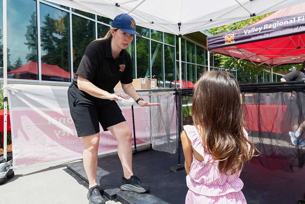 A firefighter in uniform talks to a young girl in a pink dress at an outdoor event under a tent.