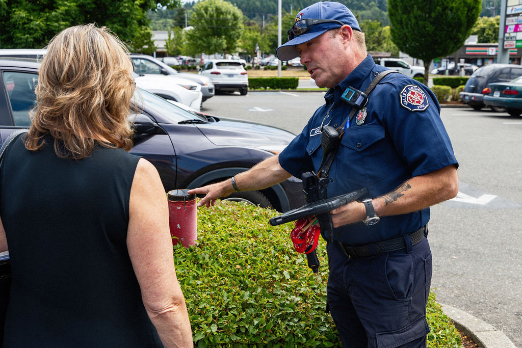 A uniformed officer talks to a woman in a parking lot, pointing at a red coffee cup placed on a hedge.