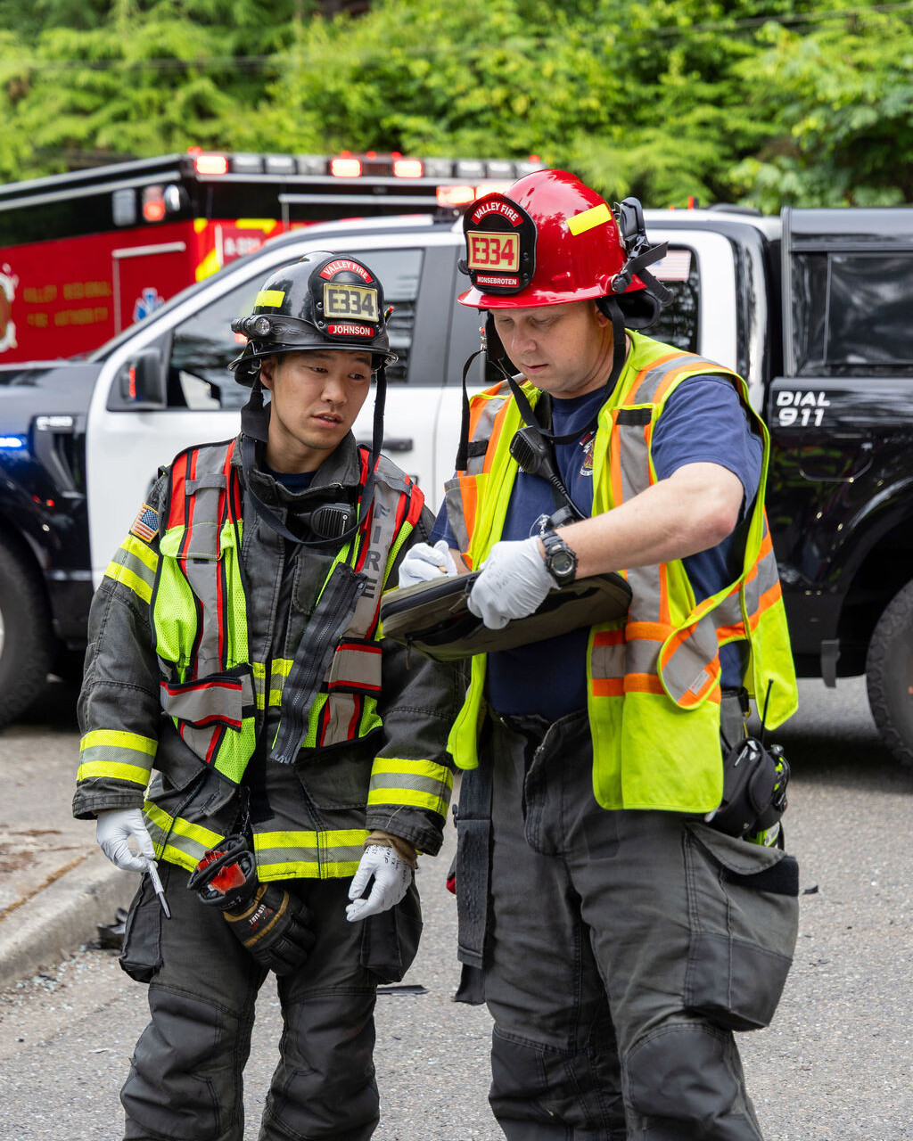 Two firefighters in uniform stand on a street; one holds a clipboard while the other listens. Emergency vehicles are parked behind them.