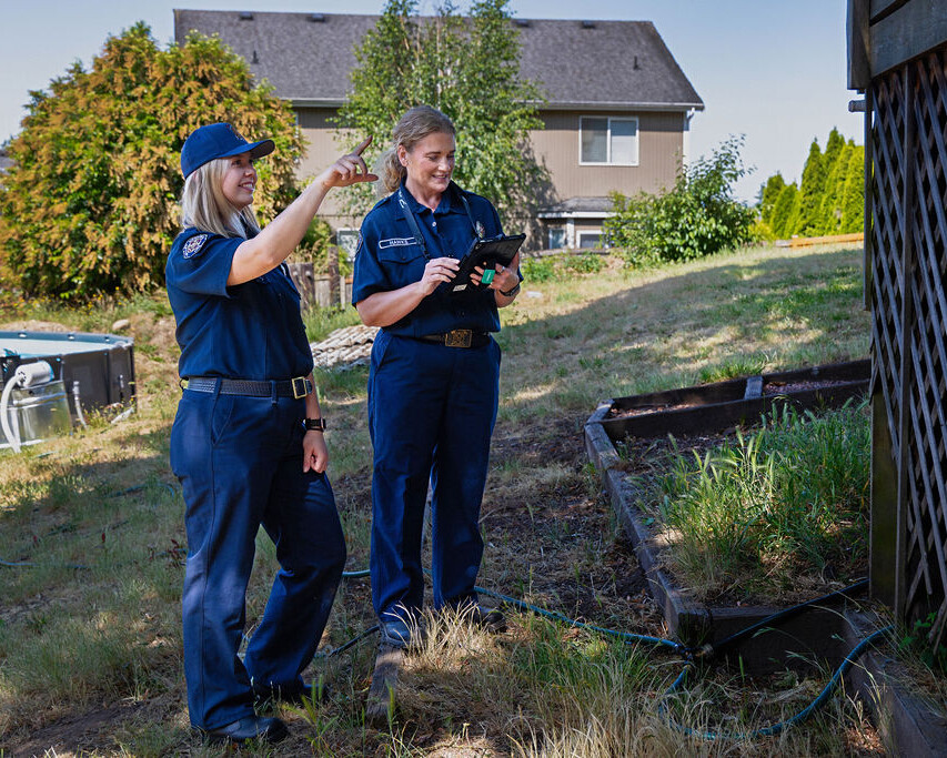 Two uniformed women stand outside a house; one points toward a structure while the other takes notes on a tablet.