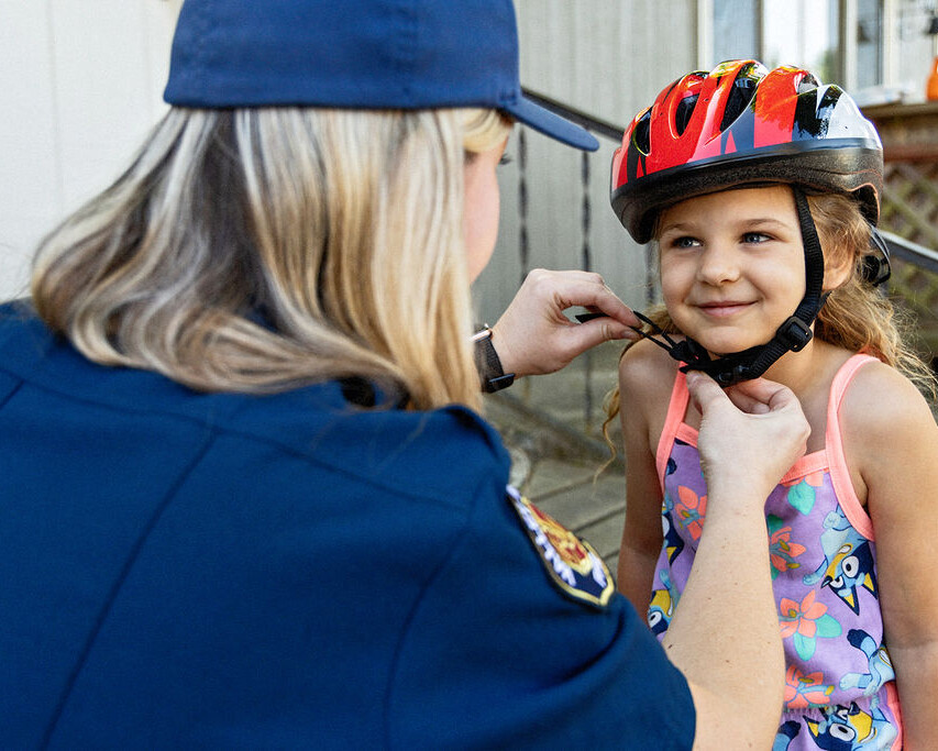 An adult in uniform adjusts the chin strap on a young girl's bicycle helmet. The girl stands outside, smiling, wearing a colorful dress.