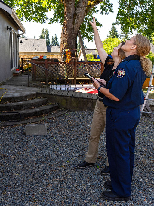 Two people, one in a fire department uniform, inspect a backyard area and look up at a tree near a house, with tools and a ladder visible in the background.