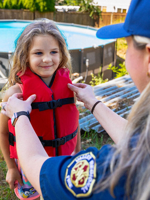 An adult helps a young girl put on a red life jacket near an above-ground swimming pool in a backyard.