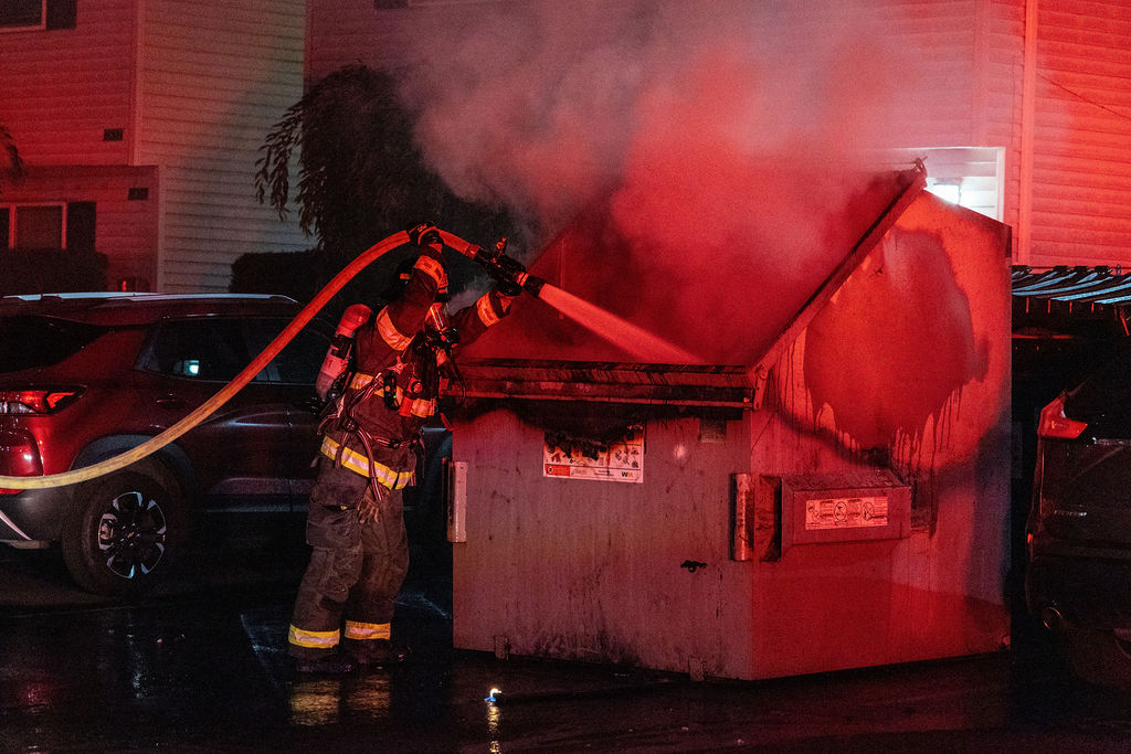 A firefighter sprays water into a smoking dumpster at night, with nearby parked cars and apartment buildings illuminated by red emergency lights.