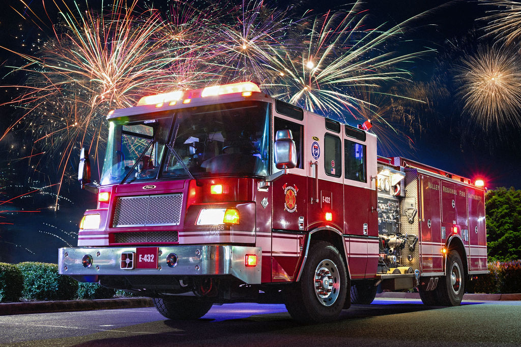 A red fire truck with lights on is parked on a street at night, with colorful fireworks bursting in the sky behind it.
