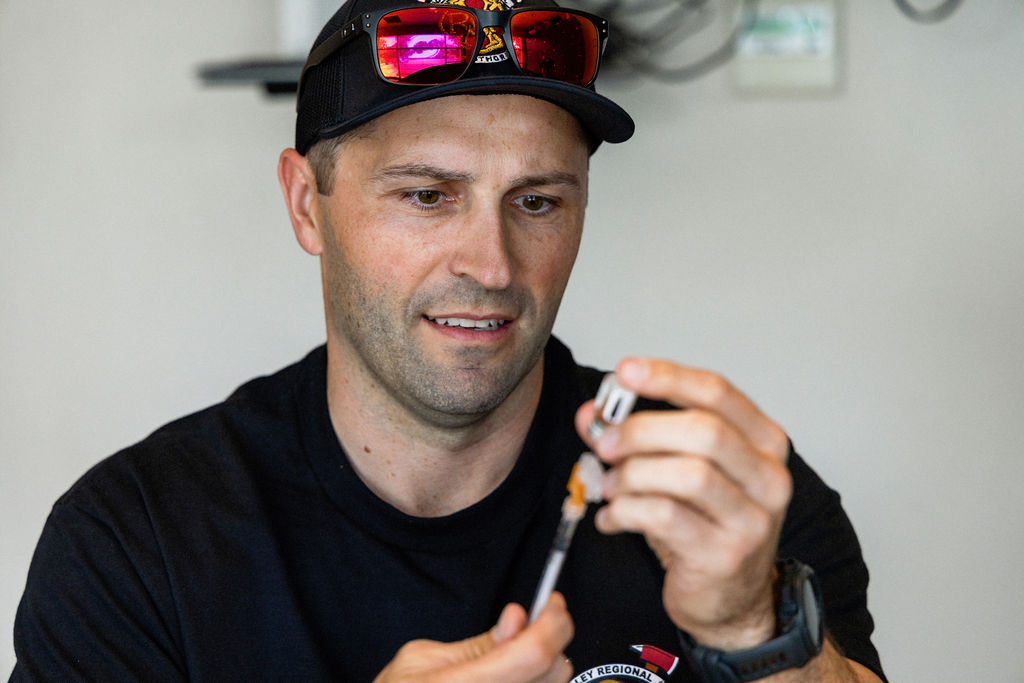 A man wearing a black cap and sunglasses prepares a syringe with medication, focusing on drawing liquid from a vial.
