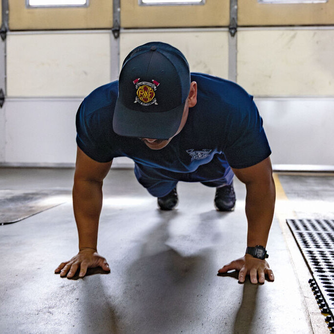 A person wearing a blue uniform and cap performs a push-up on the floor inside a garage or fire station.