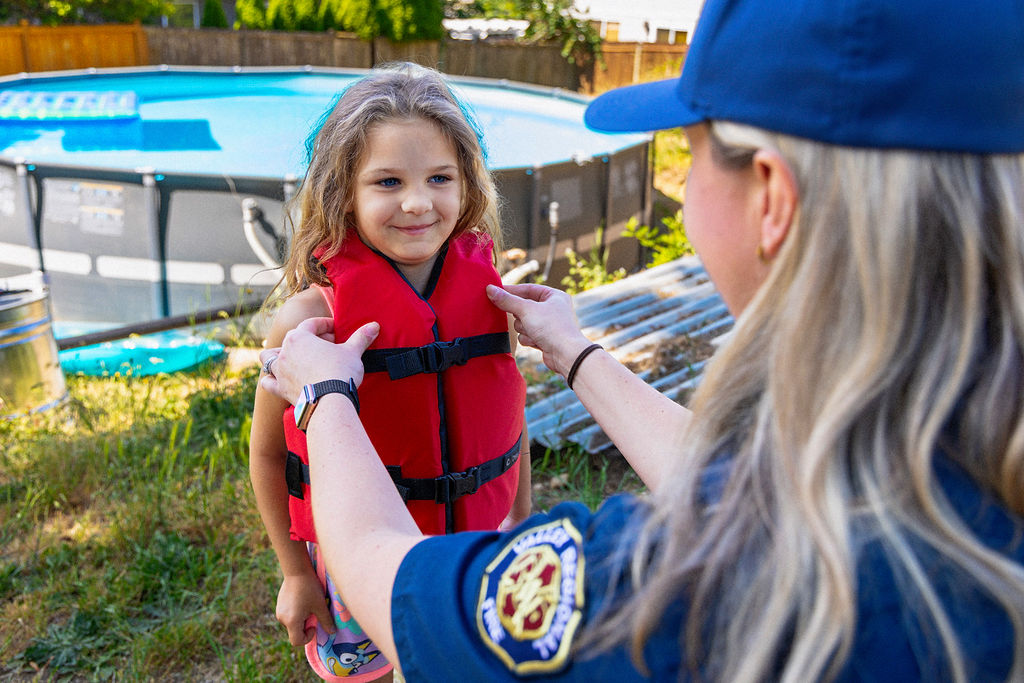 An adult helps a young girl put on a red life jacket near an above-ground pool in a backyard.