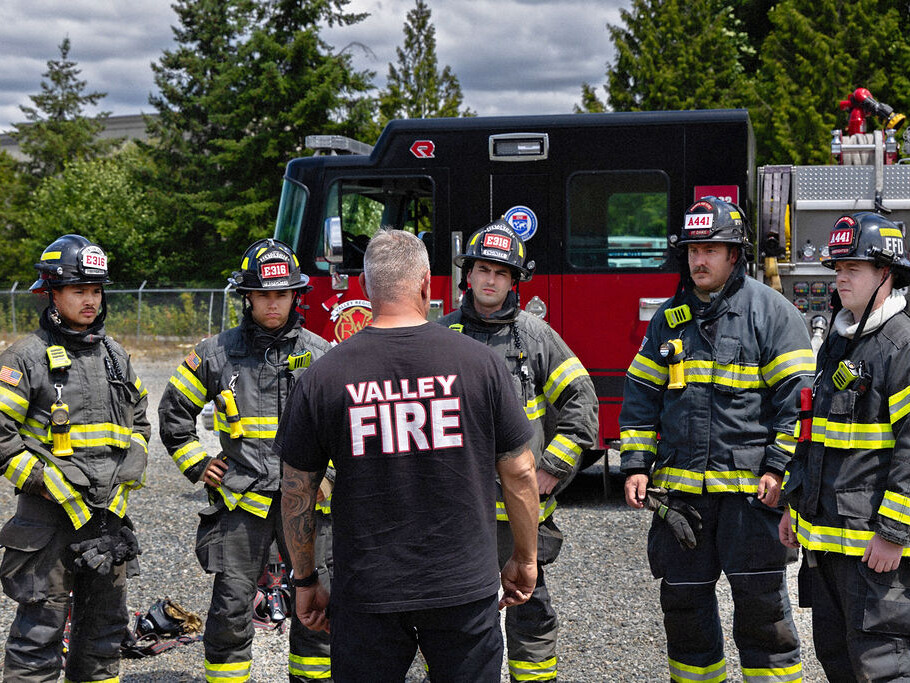 A group of firefighters in full gear listens to a briefing from a man wearing a 