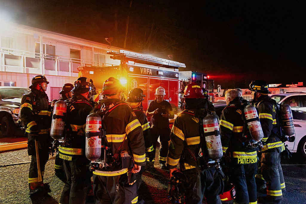 A group of firefighters in full gear stand in a circle at night near a fire truck with flashing lights in front of a building.