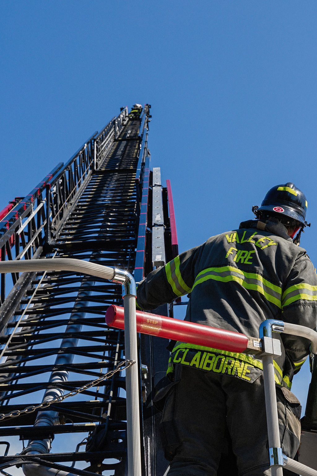 A firefighter in full gear ascends an extended ladder truck under a clear blue sky, with another firefighter visible higher up the ladder.