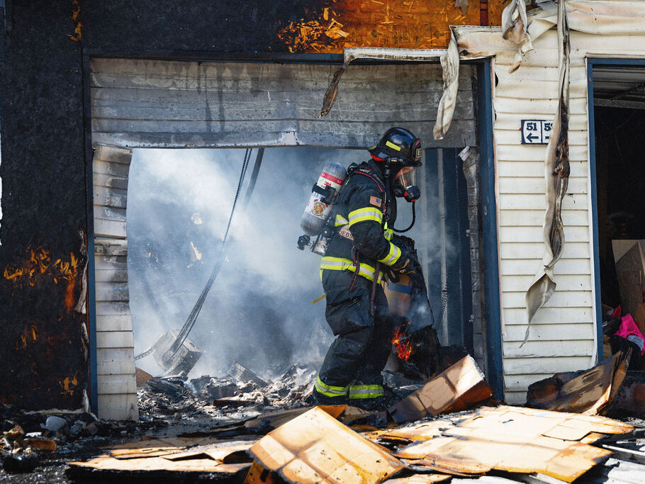 A firefighter in full gear stands in the smoke-filled entrance of a damaged garage, surrounded by debris and partially burnt boxes.