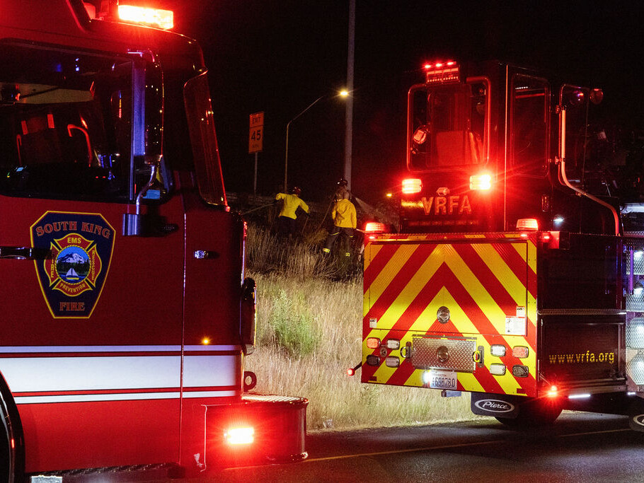 Two fire trucks with flashing lights are parked at night while firefighters in yellow jackets work nearby on a grassy roadside.