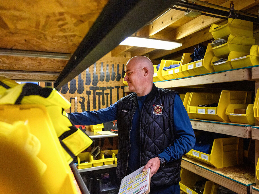 A man wearing a fire department vest organizes yellow bins and holds a checklist inside a storage room with shelves and tools.