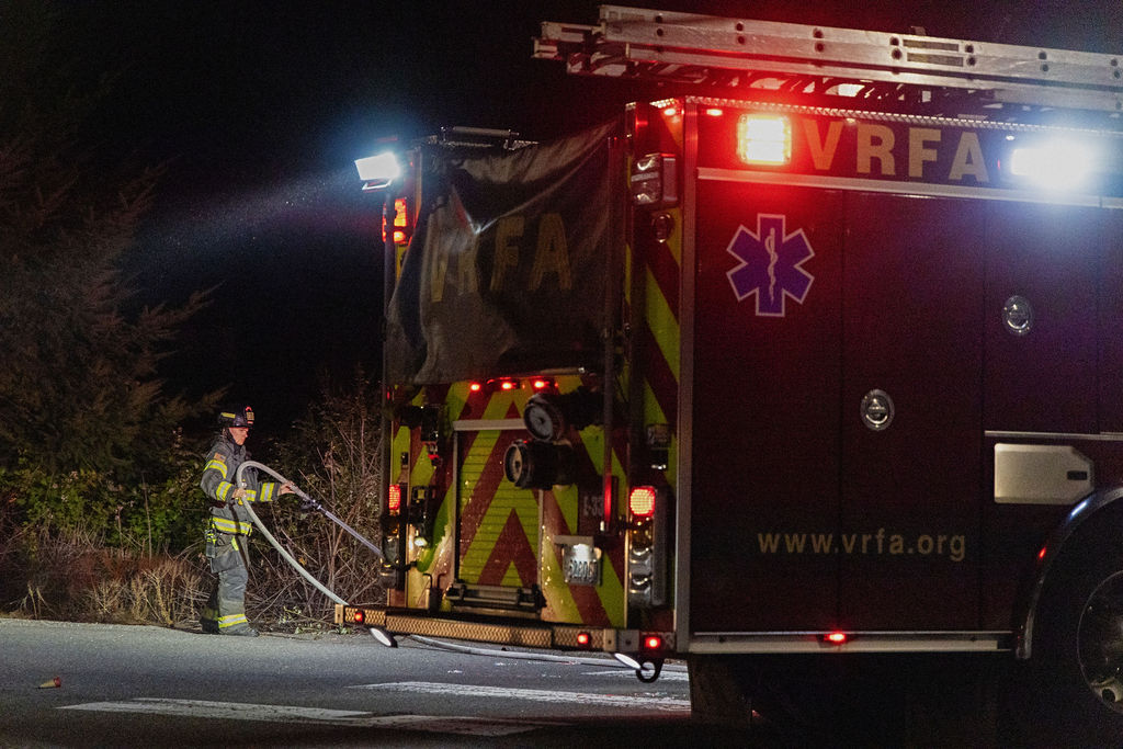 A firefighter pulls a hose from the back of a VRFA fire truck at night, with emergency lights illuminating the scene.
