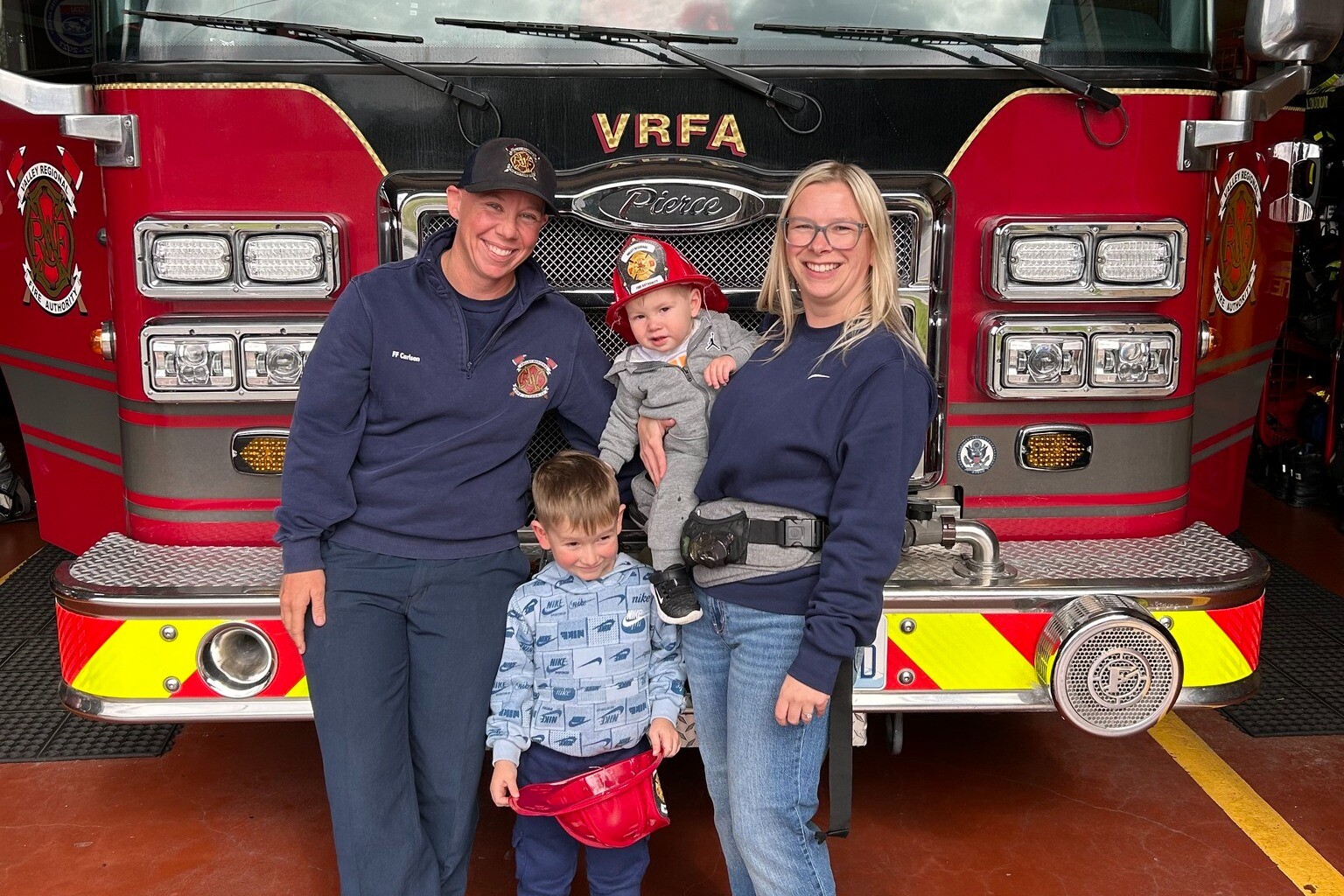 One year later, a happy reunion takes place as two adults and two children pose in front of a red fire truck inside a fire station. One adult is in uniform, while one child proudly holds a toy fire helmet.