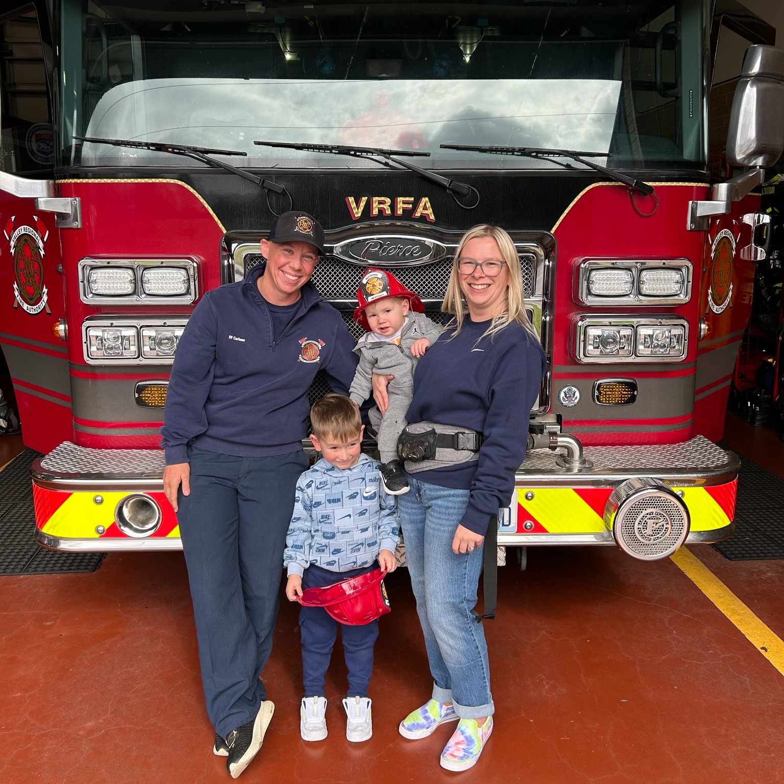 One year later, a happy reunion takes place as two adults and two children pose in front of a red fire truck inside a fire station. One adult is in uniform, while one child proudly holds a toy fire helmet.