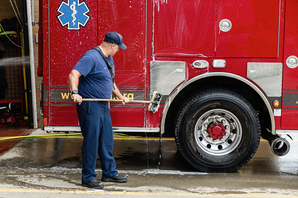 A firefighter washes the side of a red fire truck with a long brush, cleaning soap and water off the vehicle outside a station funded by the Fire Benefit Charge (FBC).