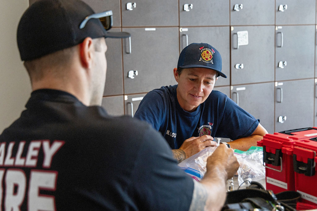 Two firefighters in uniform sit at a table with medical supplies, focusing on injury prevention safety. One faces the camera while the other has his back turned, with lockers lining the background.
