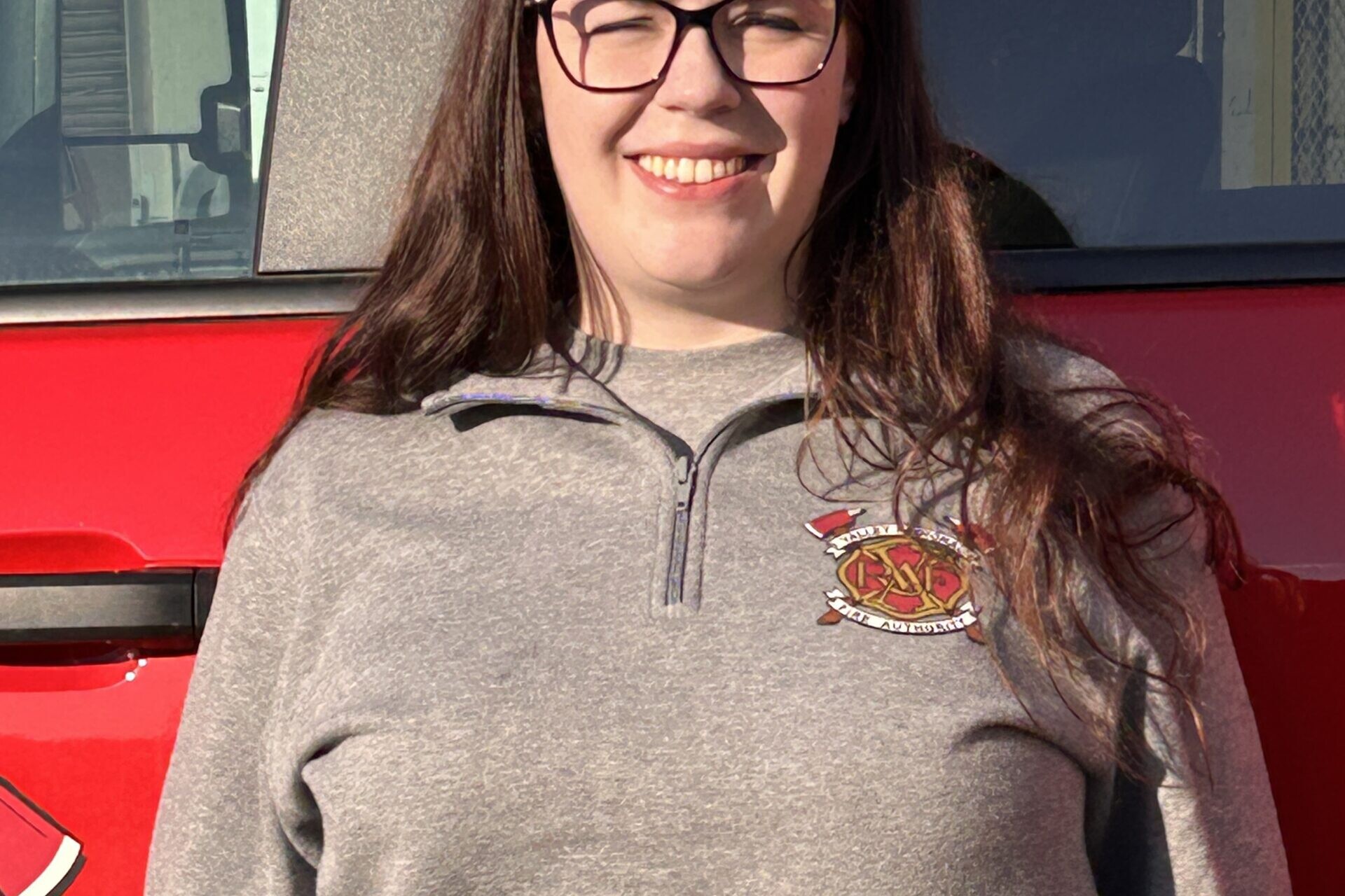 A woman with long brown hair and glasses, an intern with the VRFA CARES Team, stands in front of a red vehicle, wearing a gray pullover with an embroidered fire department emblem.