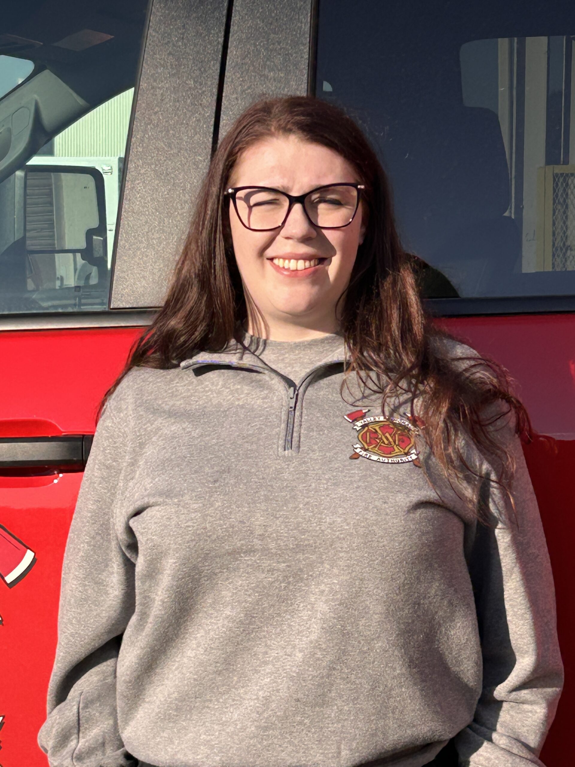 A woman with long brown hair and glasses, an intern with the VRFA CARES Team, stands in front of a red vehicle, wearing a gray pullover with an embroidered fire department emblem.