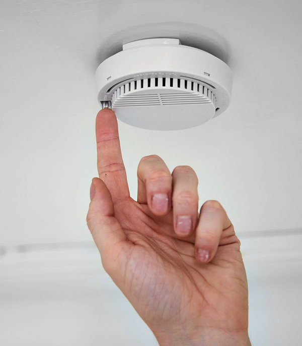 A hand presses the test button on a ceiling-mounted smoke detector, ensuring Fire Alarms work properly.