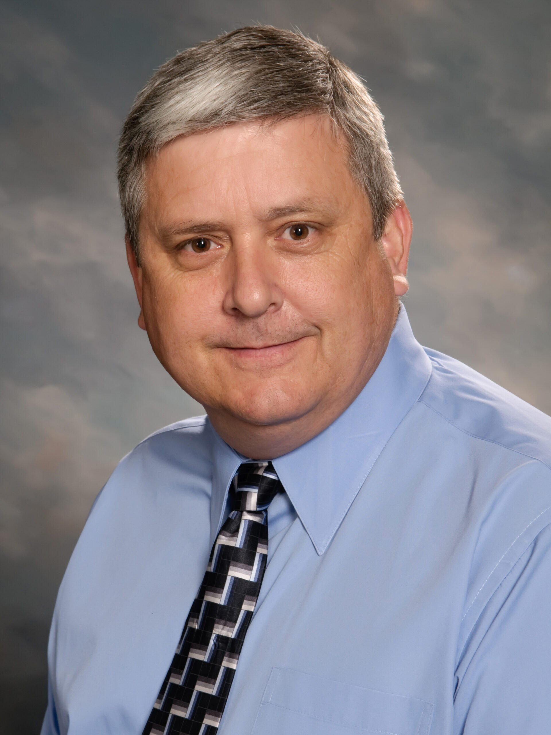 A middle-aged man with short gray hair wearing a light blue dress shirt and a patterned tie, posing against a neutral studio background.