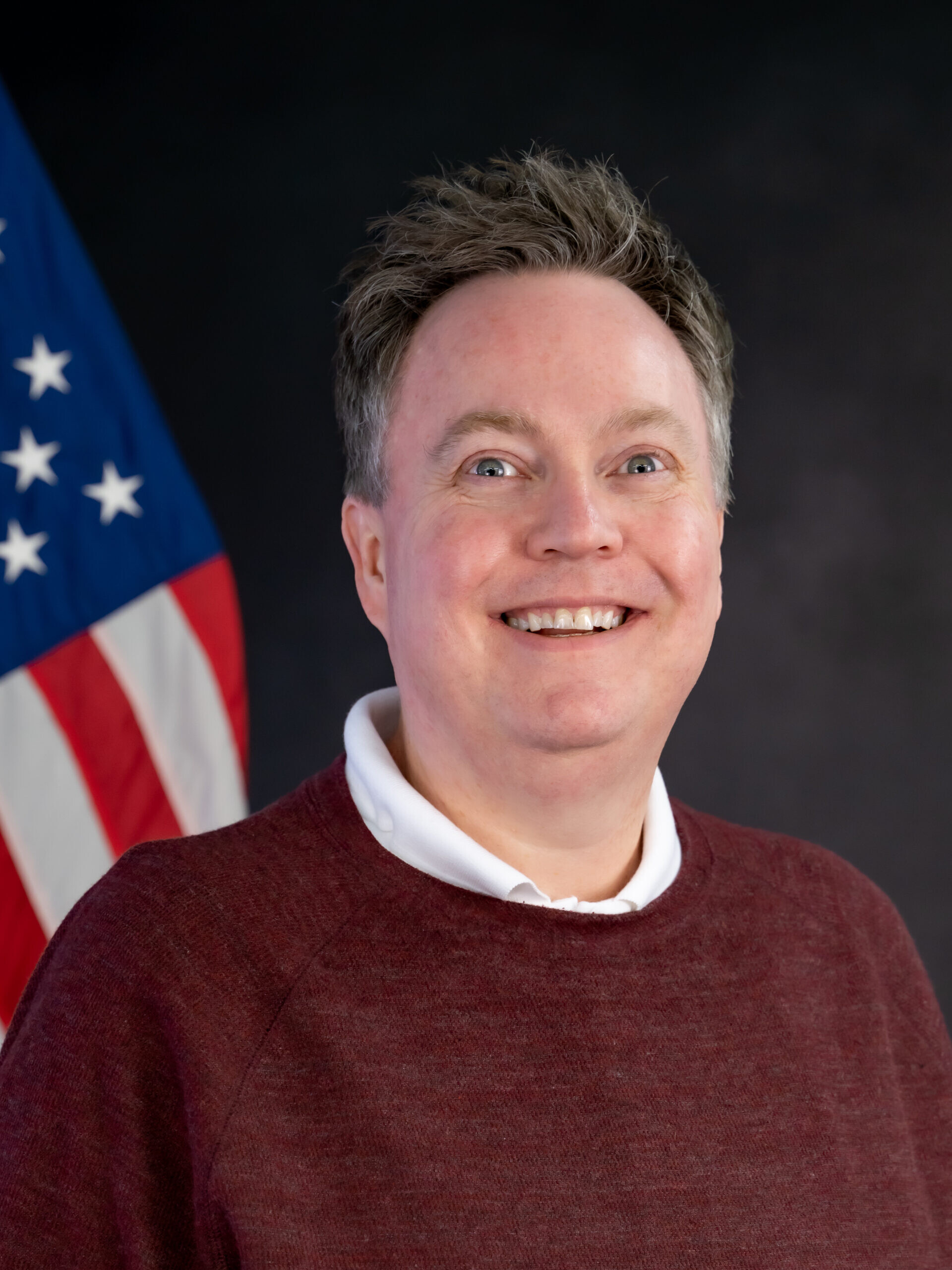 A person in a maroon sweater and white collared shirt smiles in front of a U.S. flag and a dark background.