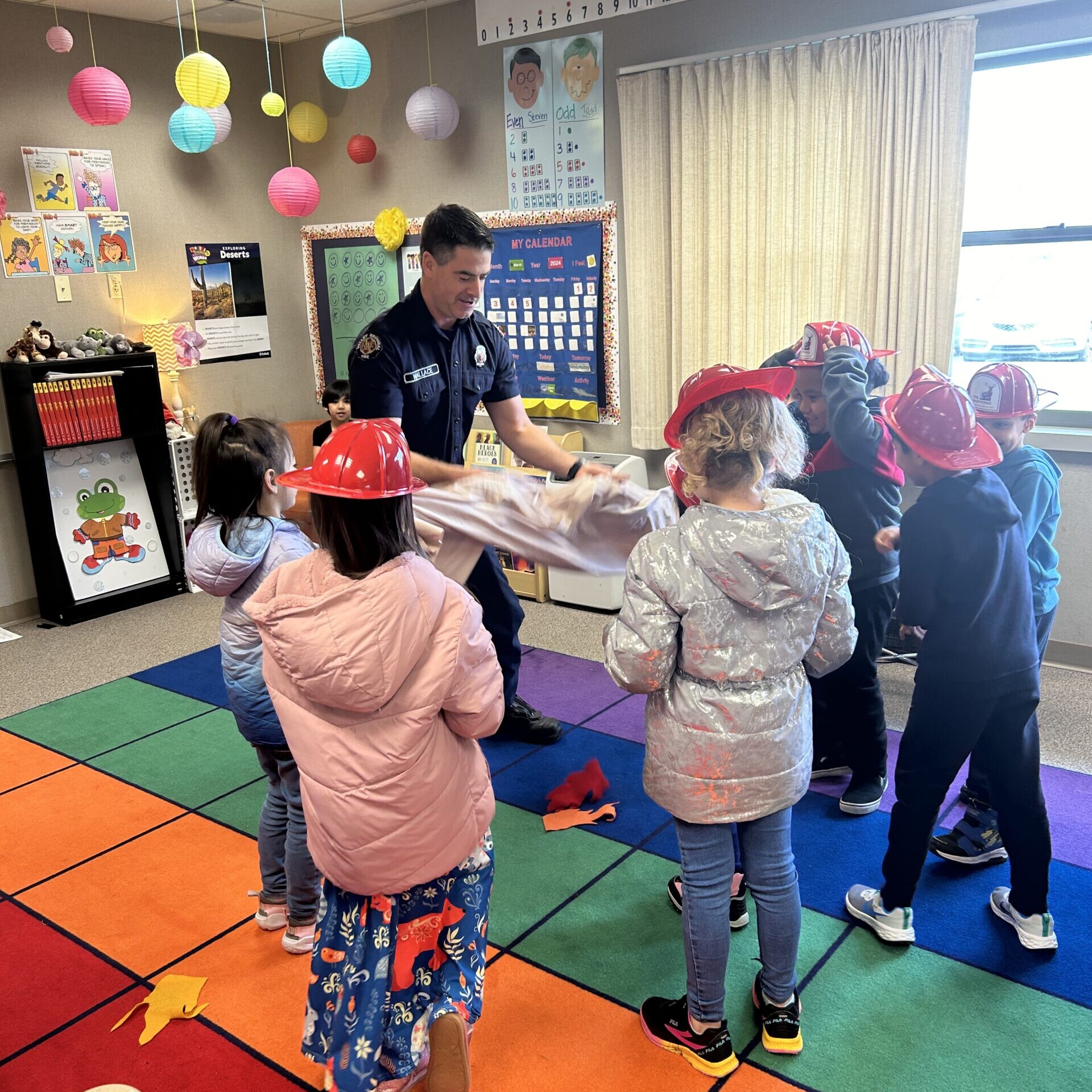 A group of children wearing toy firefighter helmets gathers around an adult in uniform demonstrating Home Fire Prevention with a blanket in a colorful classroom.