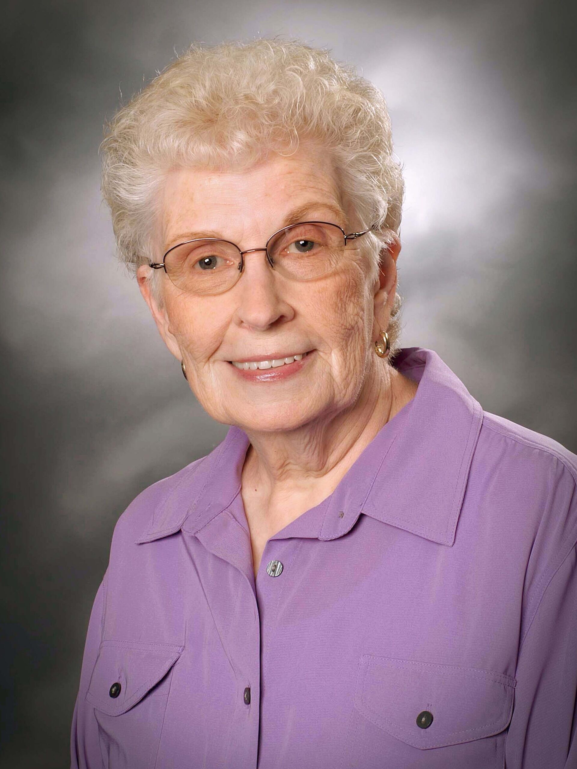 Elderly woman with short, curly gray hair, glasses, and a light purple collared shirt, smiling in front of a gray studio backdrop.