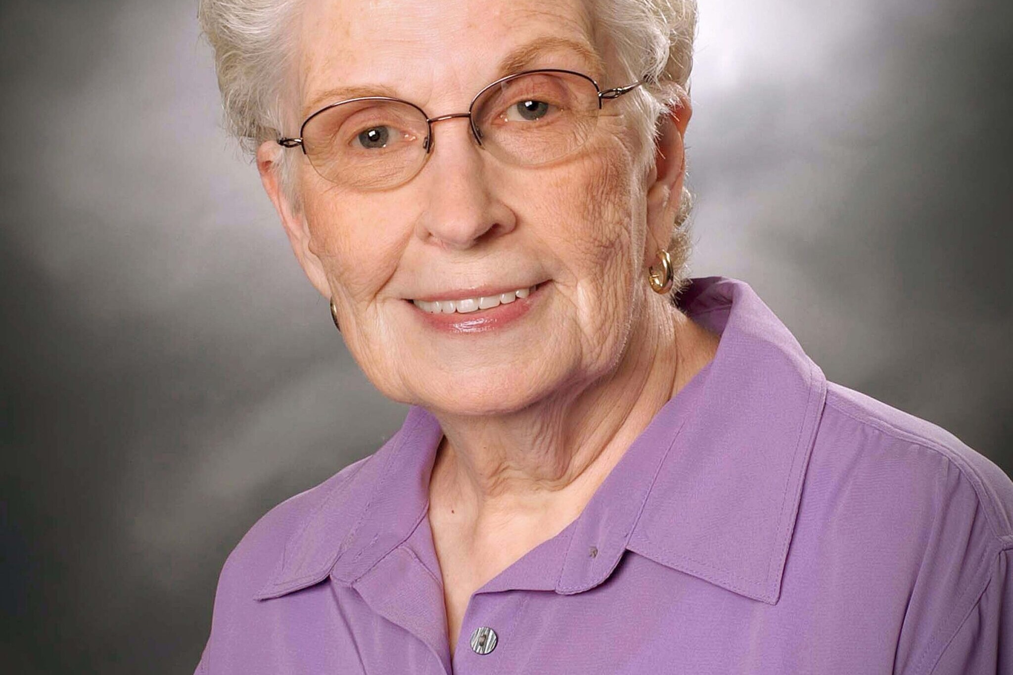 Elderly woman with short, curly gray hair, glasses, and a light purple collared shirt, smiling in front of a gray studio backdrop.