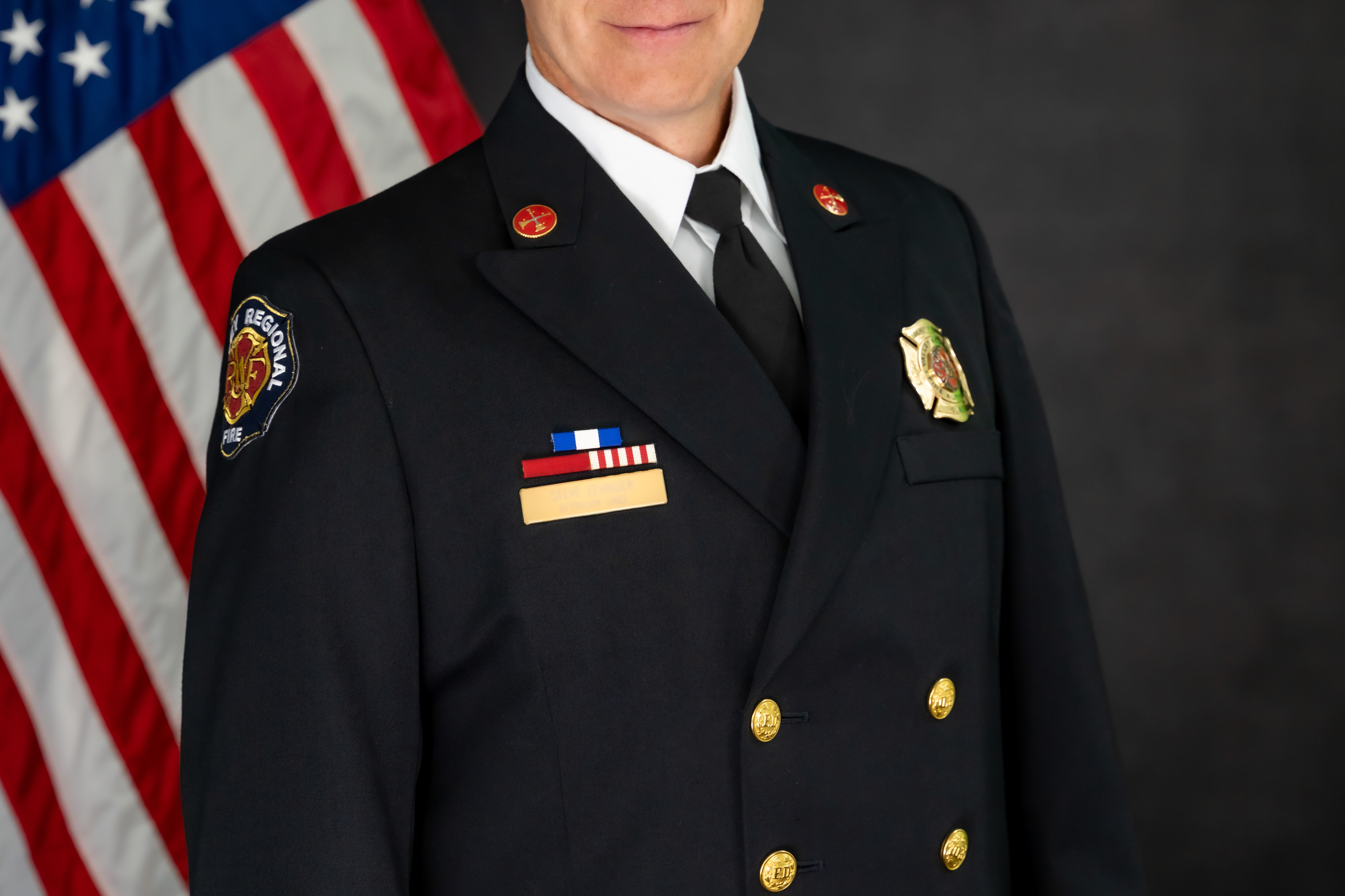 A uniformed fire department officer stands in front of a U.S. flag, facing the camera with a neutral expression, marking Battalion Chief retirement and honoring years of dedicated service.