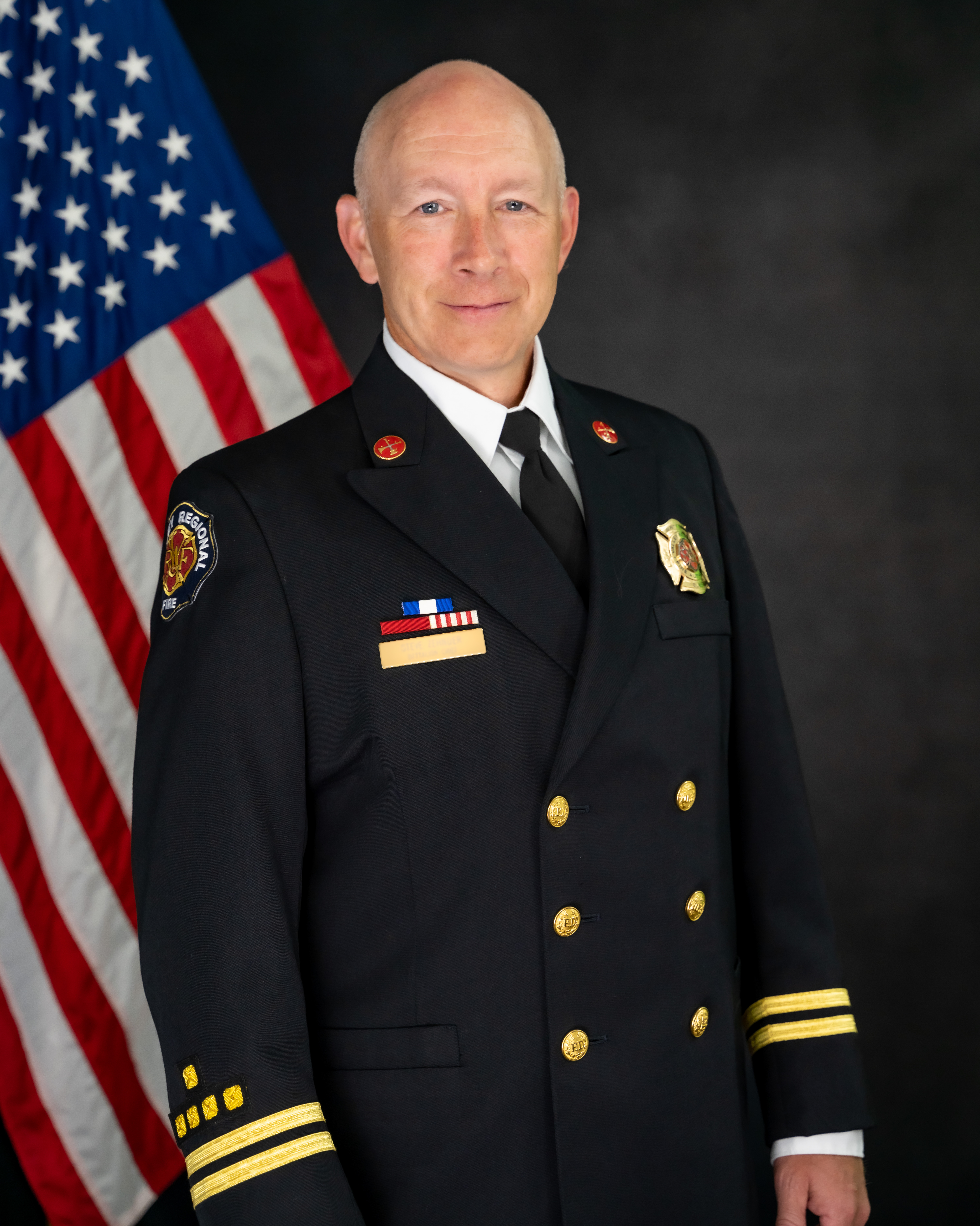 A uniformed fire department officer stands in front of a U.S. flag, facing the camera with a neutral expression, marking Battalion Chief retirement and honoring years of dedicated service.
