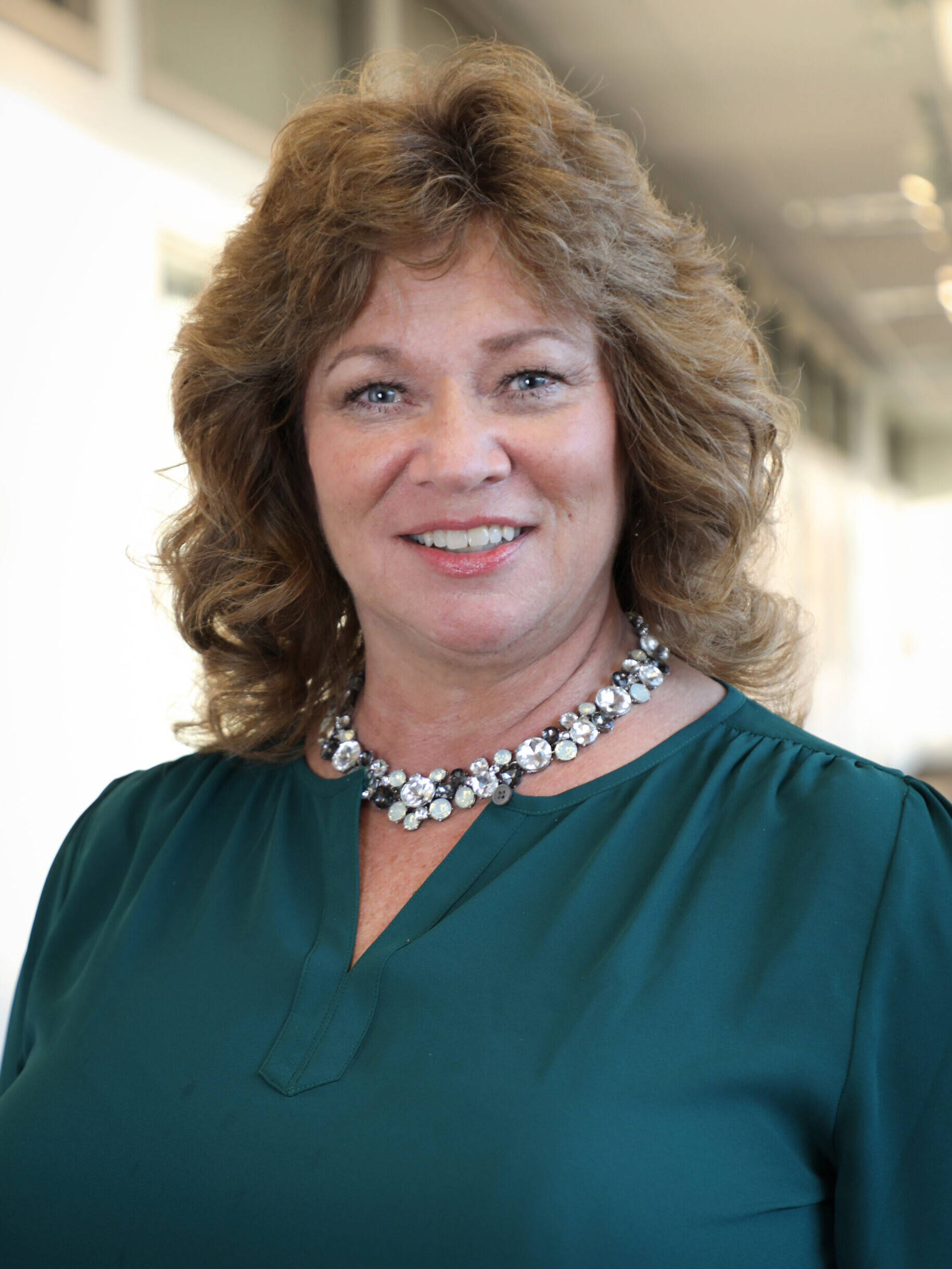 A woman with curly brown hair, wearing a green top and a jeweled necklace, smiles while standing indoors in a well-lit hallway.