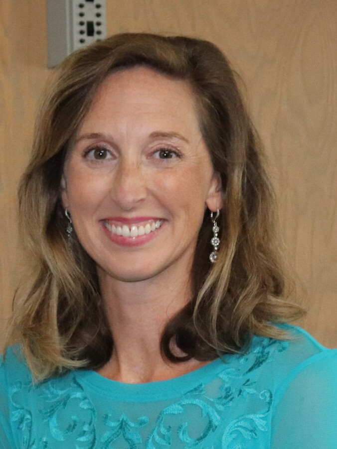 A woman with shoulder-length brown hair, wearing a turquoise top and dangling earrings, smiles in front of a wooden door.