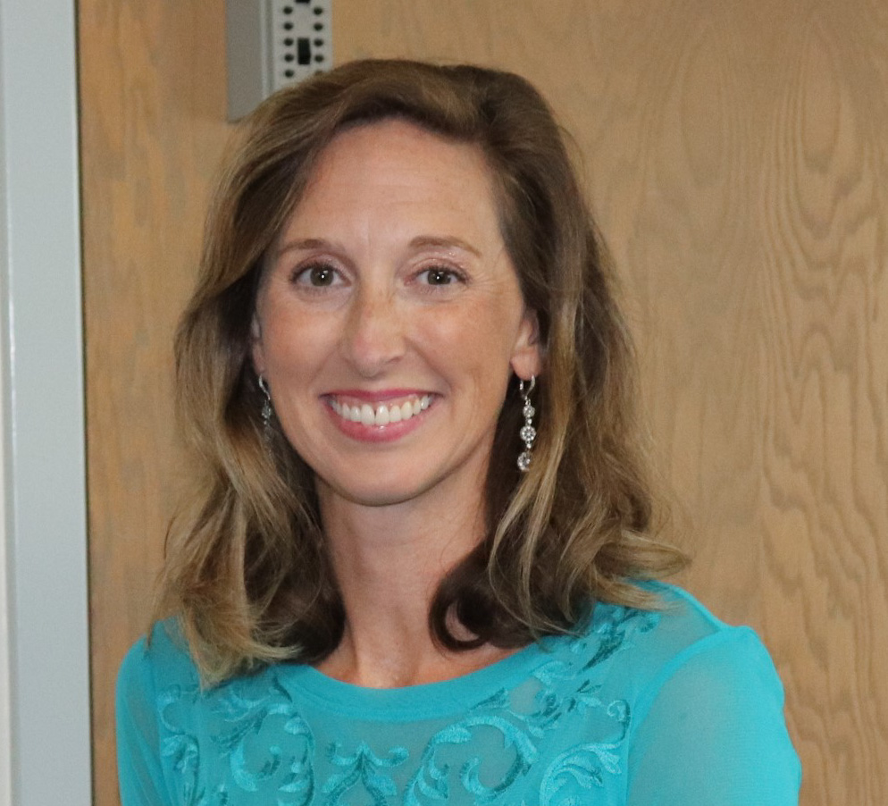 A woman with shoulder-length brown hair, wearing a turquoise top and dangling earrings, smiles in front of a wooden door.
