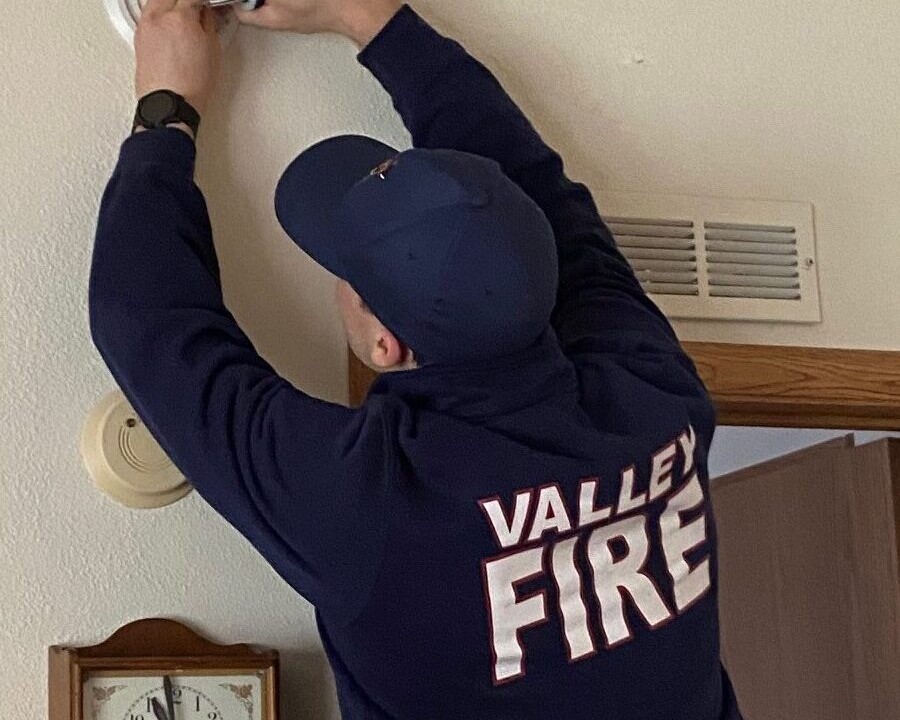 A firefighter in uniform installs or checks a smoke detector on a wall above a clock and next to a door inside a house, providing important safety information for the household.