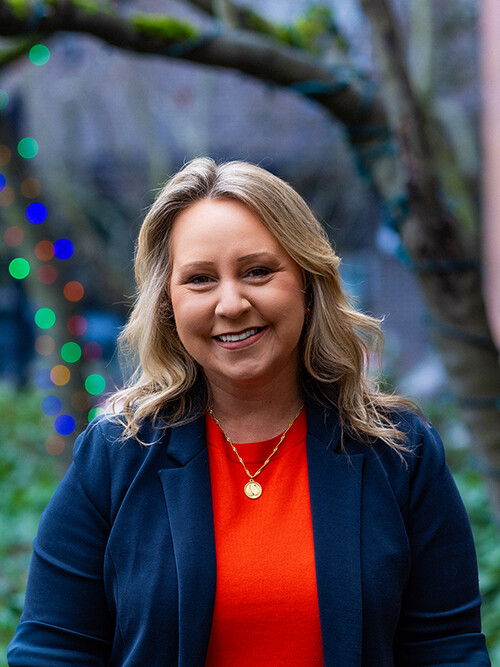 Woman with blonde hair wearing a blue blazer and orange top stands outdoors, smiling. Blurred trees and colorful lights are in the background.