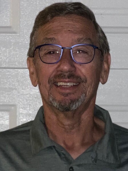 A man with short gray hair, glasses, and a goatee is smiling at the camera, wearing a gray polo shirt and standing in front of a white textured wall.