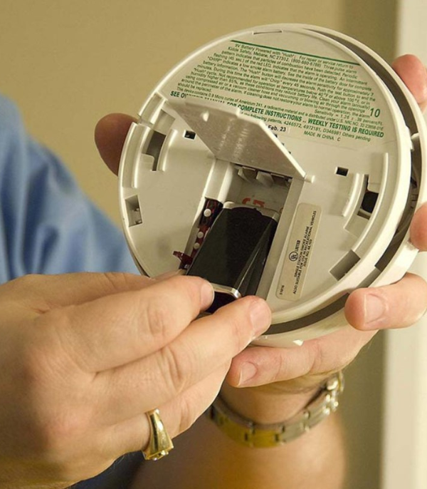 A person replaces the battery in a smoke detector, holding the device open to insert a new 9-volt battery, ensuring fire alarms remain operational.