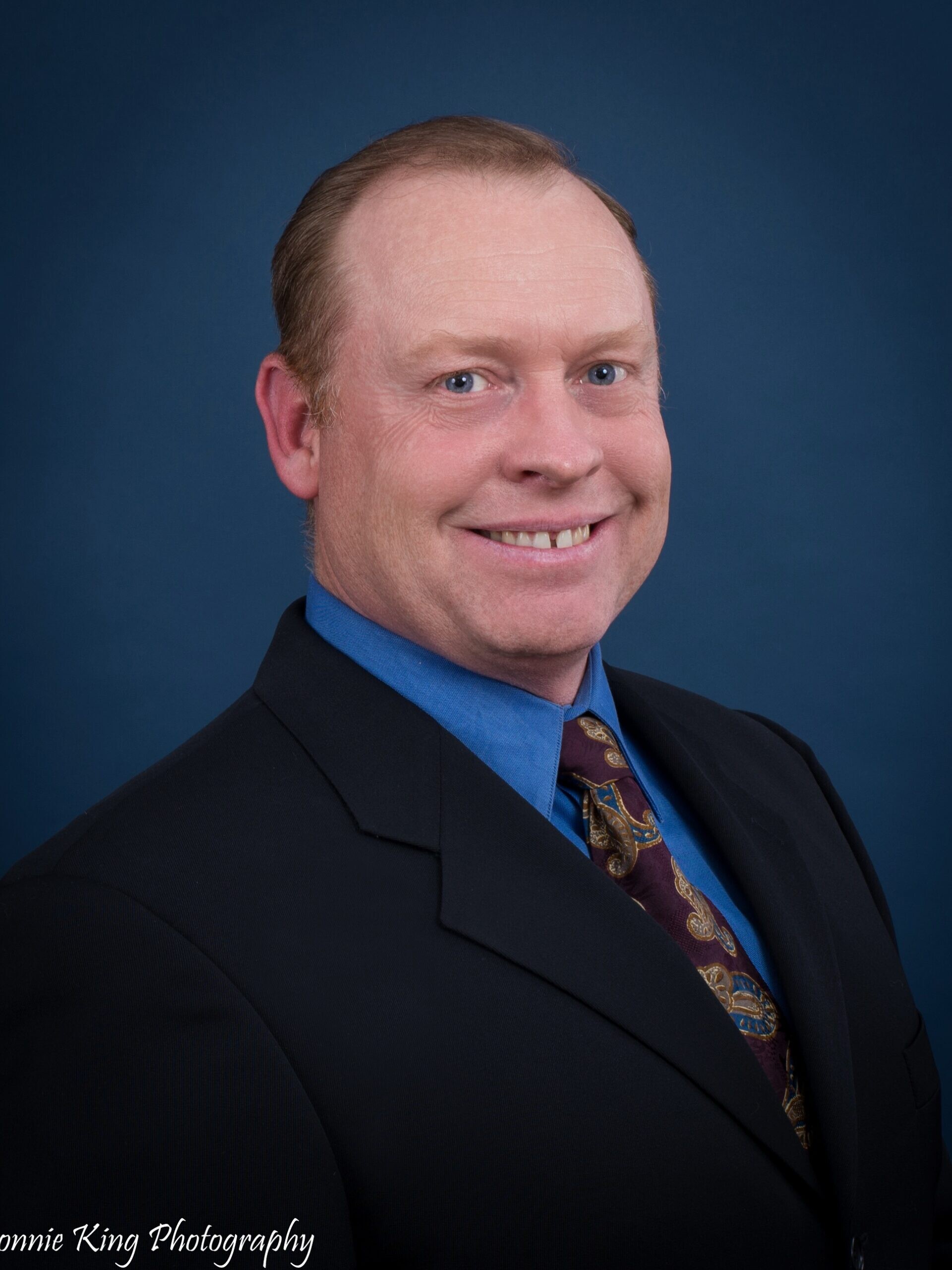 A man in a suit and tie smiles for a professional studio portrait against a dark blue background.