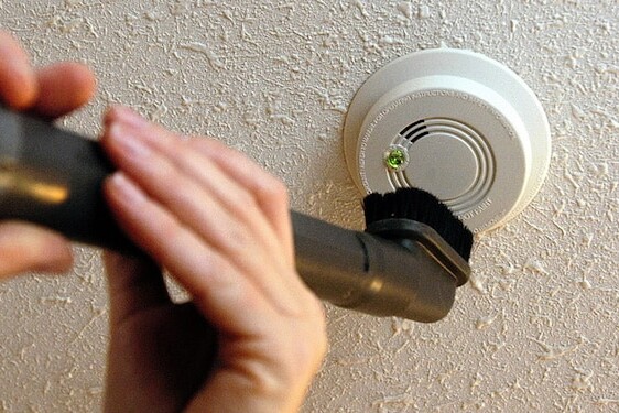 A person uses a vacuum cleaner with a brush attachment to clean a ceiling-mounted fire alarm.