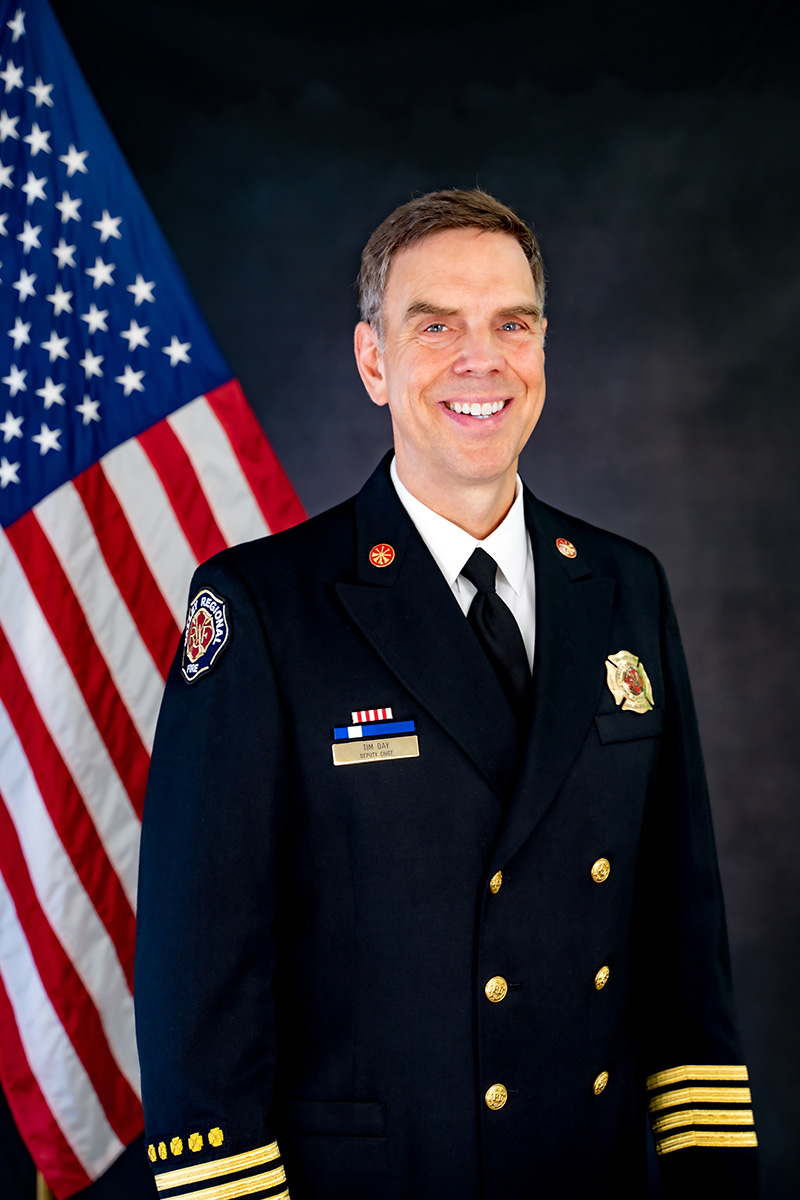 A man in a formal fire department uniform, symbolizing Governance Administration, stands smiling in front of a U.S. flag and a dark background.