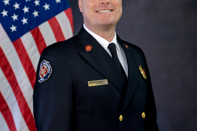 A man in a formal fire department uniform, representing Governance Administration, stands in front of a U.S. flag, smiling at the camera against a dark studio background.