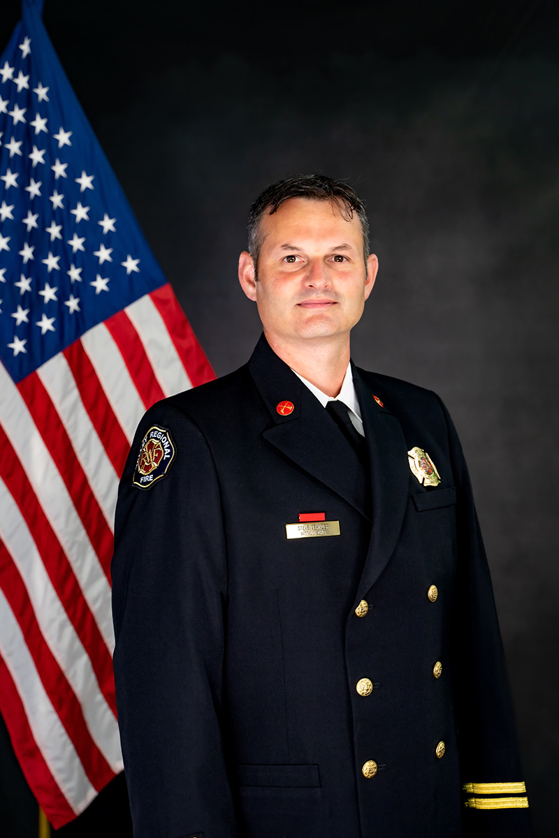 A man in a formal firefighter uniform stands before an American flag, representing Governance Administration as he poses for a professional portrait against a dark background.