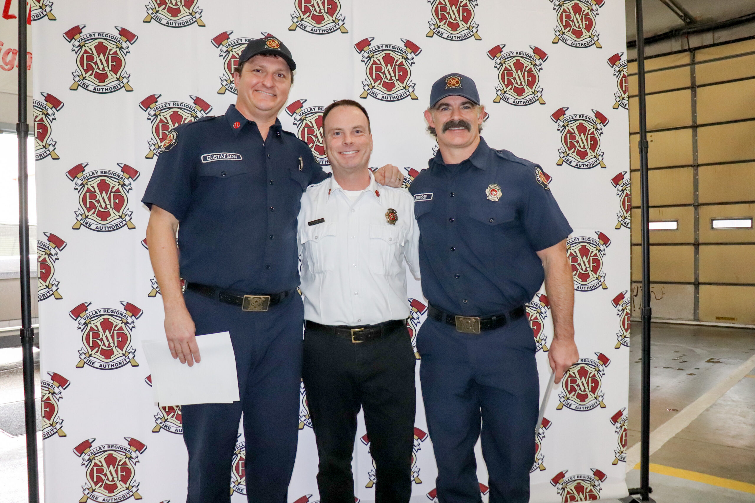 Three firefighters in uniform stand together indoors, smiling, in front of a banner with fire department logos, after receiving the Medal of Merit for a heroic rescue led by Captain Jordan Gustafson.