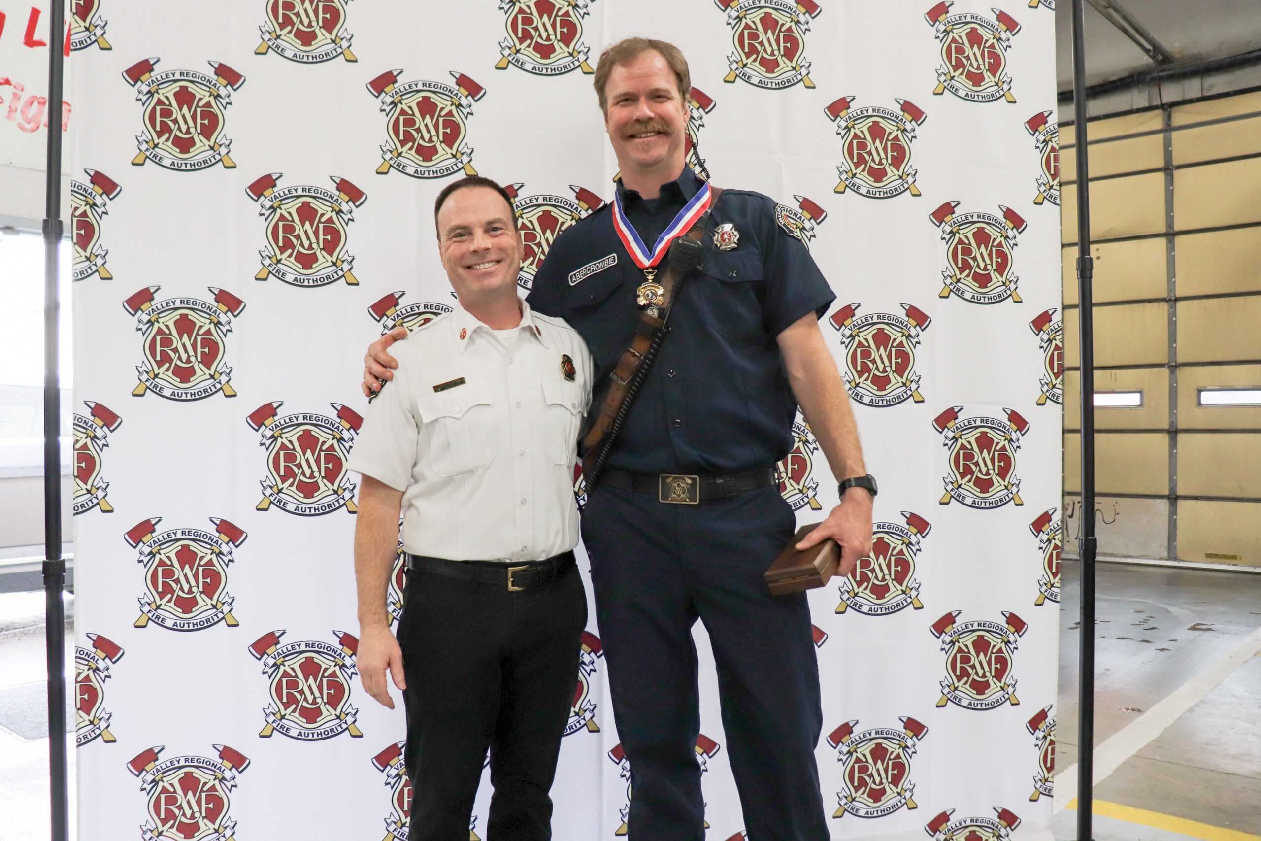 Two firefighters stand smiling in front of a backdrop with fire department logos; Firefighter John Abercrombie wears a Medal of Honor and holds a plaque, suggesting an award ceremony.