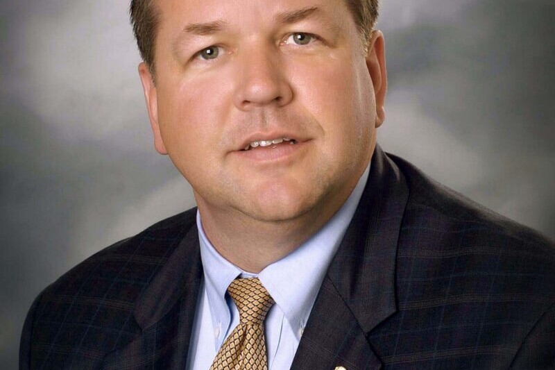 Eric Robertson, the founding administrator of Valley Regional Fire Authority, poses in a suit and tie for a formal portrait against a neutral, cloudy background.