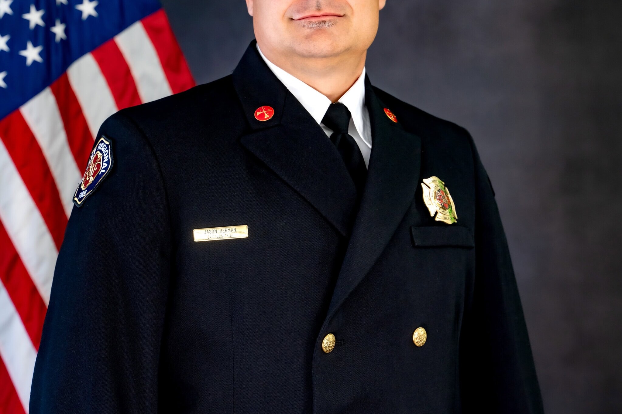 A man in a formal fire department dress uniform stands in front of an American flag, facing the camera with a neutral expression, exemplifying the professionalism and dedication essential to effective Governance Administration.