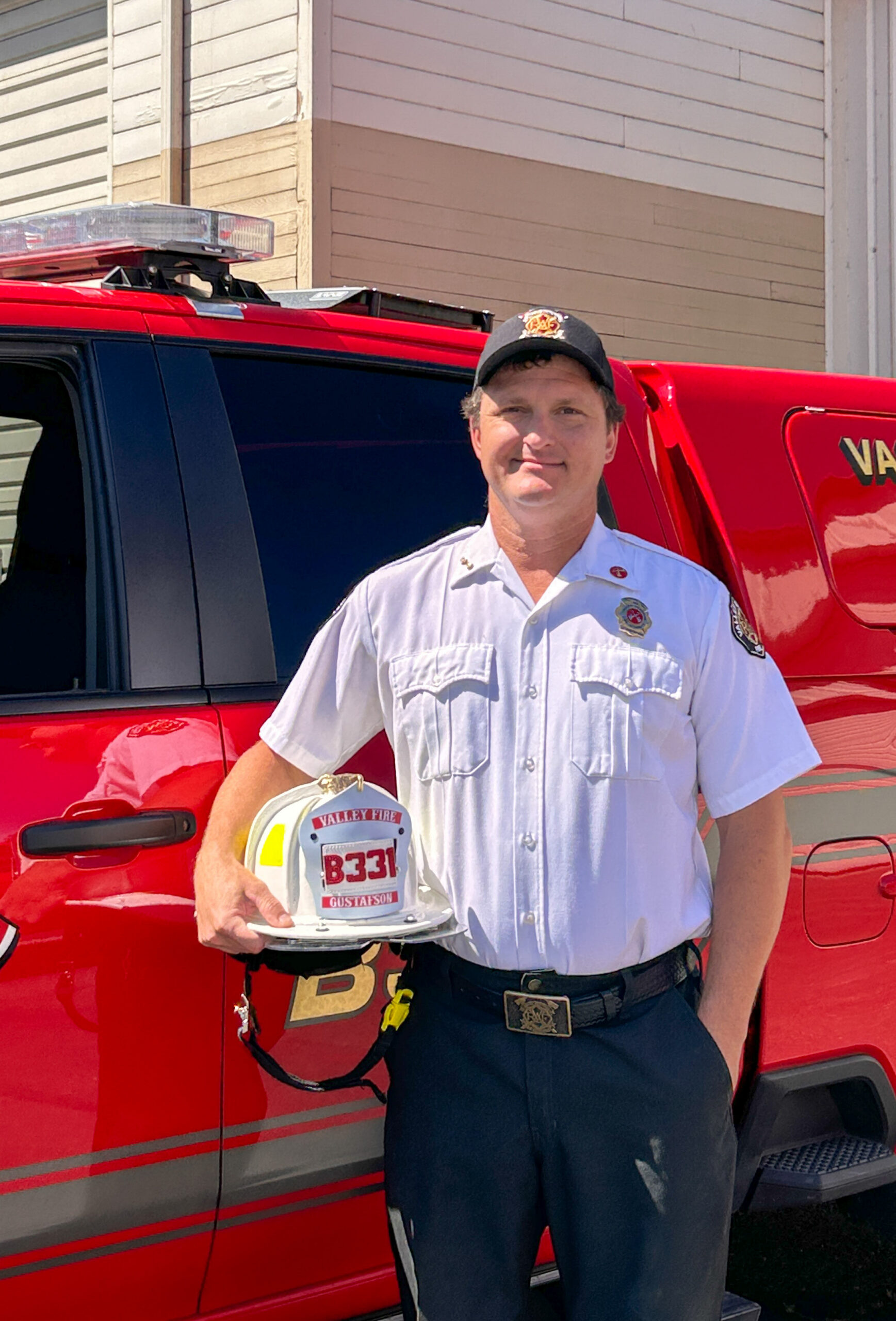 A firefighter in uniform stands next to a red emergency vehicle, holding a white helmet labeled
